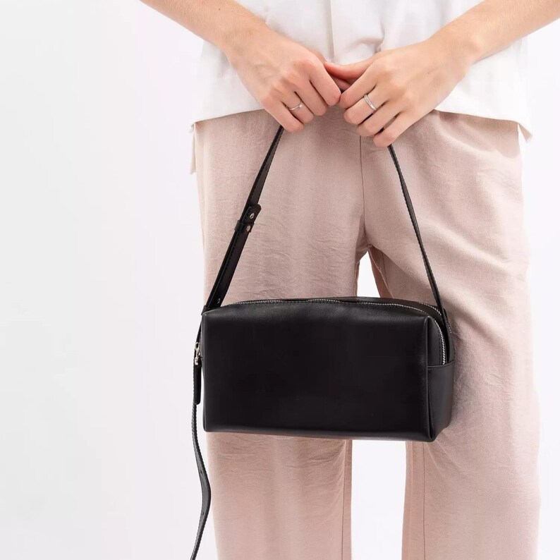 May include: A rectangular black shoulder bag with a smooth finish, featuring a top zip closure and a thin, adjustable shoulder strap. The bag has a structured, boxy silhouette with clean lines and silver-toned hardware accents on the strap.