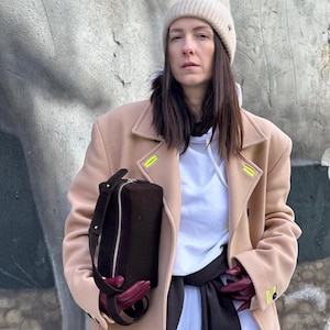 Pu&ograve; includere: Una borsa a tracolla rettangolare marrone scuro con cerniera argentata, indossata con un cappotto di lana beige con accenti giallo neon, una felpa bianca con cappuccio, un berretto in maglia beige e guanti in pelle bordeaux scuro.