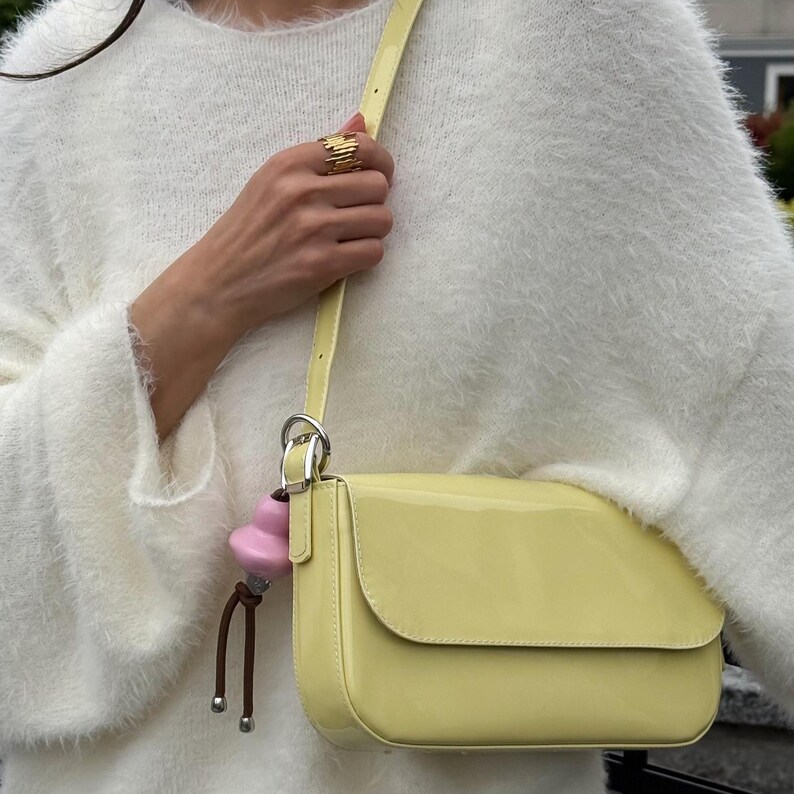 May include: A pale yellow patent leather shoulder bag with a flap closure and a matching adjustable strap. A silver-toned metal ring attaches a small pink gourd-shaped charm with two brown cords and silver beads to the side of the bag.
