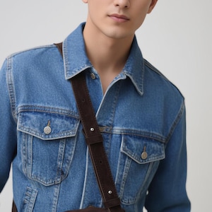 Pu&ograve; includere: Una giacca in denim blu con colletto classico, due tasche sul petto con chiusura a bottone e maniche lunghe, indossata con una borsa a tracolla in camoscio marrone scuro con tracolla regolabile e chiusura a zip in metallo argentato.