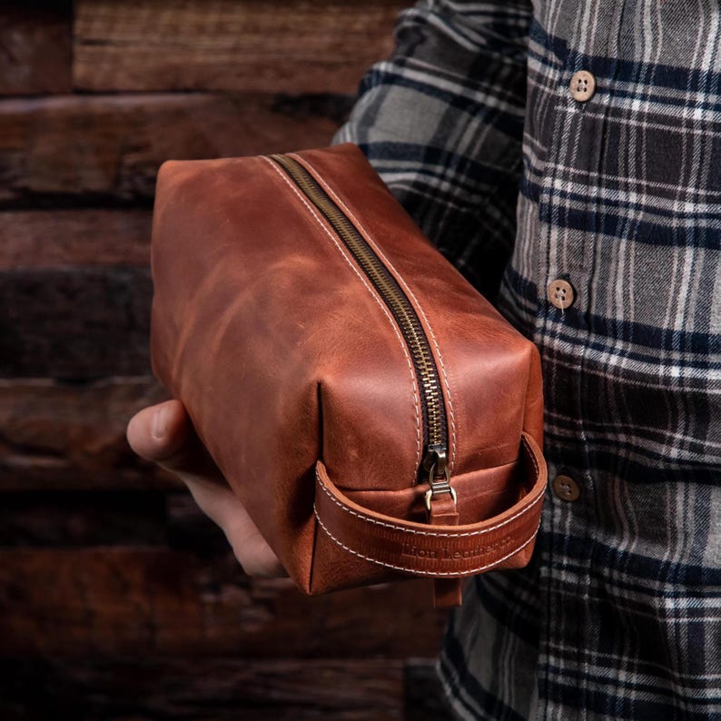May include: A rectangular brown leather wash bag featuring a brass-toned metal zip closure running along the top and a matching leather side handle with white contrast stitching. The bag has a smooth, matte finish and a structured shape.