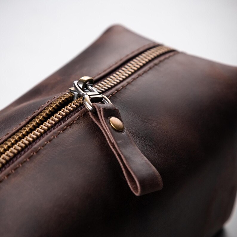 May include: A dark brown leather toiletry bag featuring a sturdy brass-toned metal zip closure. The zip pull is constructed from a matching leather tab secured with a small, circular brass-coloured metal rivet, providing a functional grip.