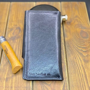 Puede incluir: Una cartera de cuero negro con orientaci&oacute;n vertical y costuras visibles a lo largo de los bordes. El texto Lion Leather co. est&aacute; grabado en la esquina inferior. Una navaja plegable con mango de madera se sit&uacute;a a la izquierda de la cartera.