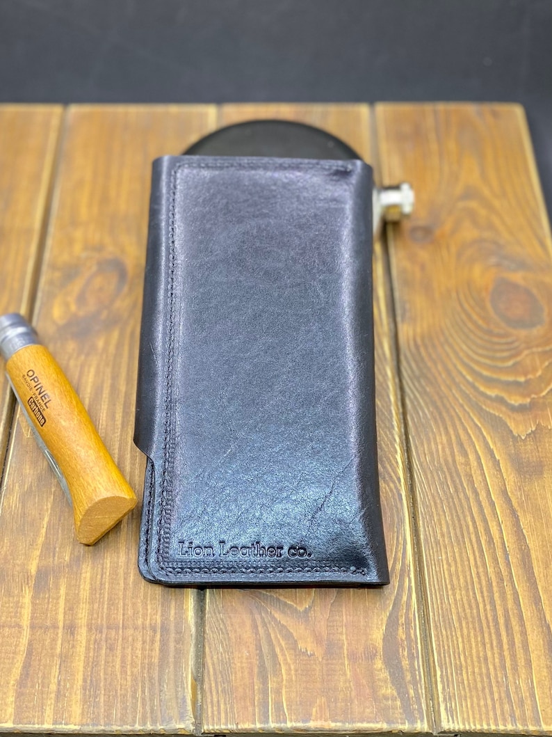 Puede incluir: Una cartera de cuero negro con orientaci&oacute;n vertical y costuras visibles a lo largo de los bordes. El texto Lion Leather co. est&aacute; grabado en la esquina inferior. Una navaja plegable con mango de madera se sit&uacute;a a la izquierda de la cartera.