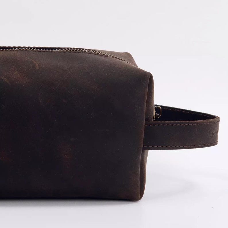 May include: A dark brown leather wash bag with a matte finish and a visible grain texture. The rectangular pouch features a sturdy side handle made of matching brown leather and a dark metal zip closure running along the top edge.