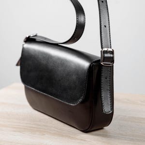 Op de afbeelding: Een zwarte leren schoudertas met een gladde voorflap, een afgeronde onderkant en een verstelbare riem met een zilverkleurige metalen gesp. De tas heeft een gestructureerd silhouet met strakke stiksels langs de randen en een glanzende afwerking.