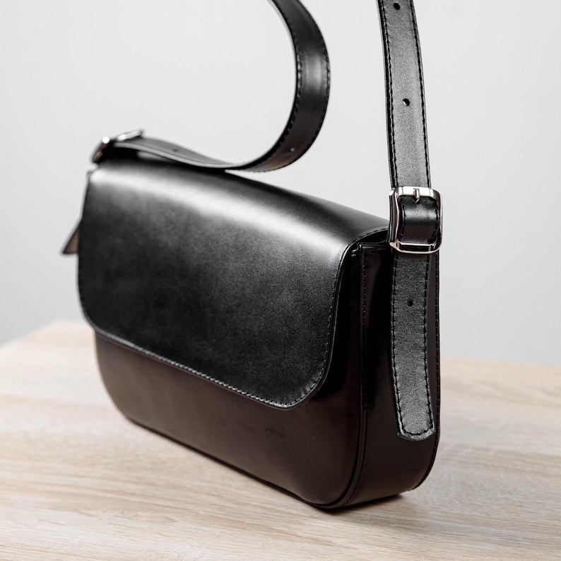 Op de afbeelding: Een zwarte leren schoudertas met een gladde voorflap, een afgeronde onderkant en een verstelbare riem met een zilverkleurige metalen gesp. De tas heeft een gestructureerd silhouet met strakke stiksels langs de randen en een glanzende afwerking.