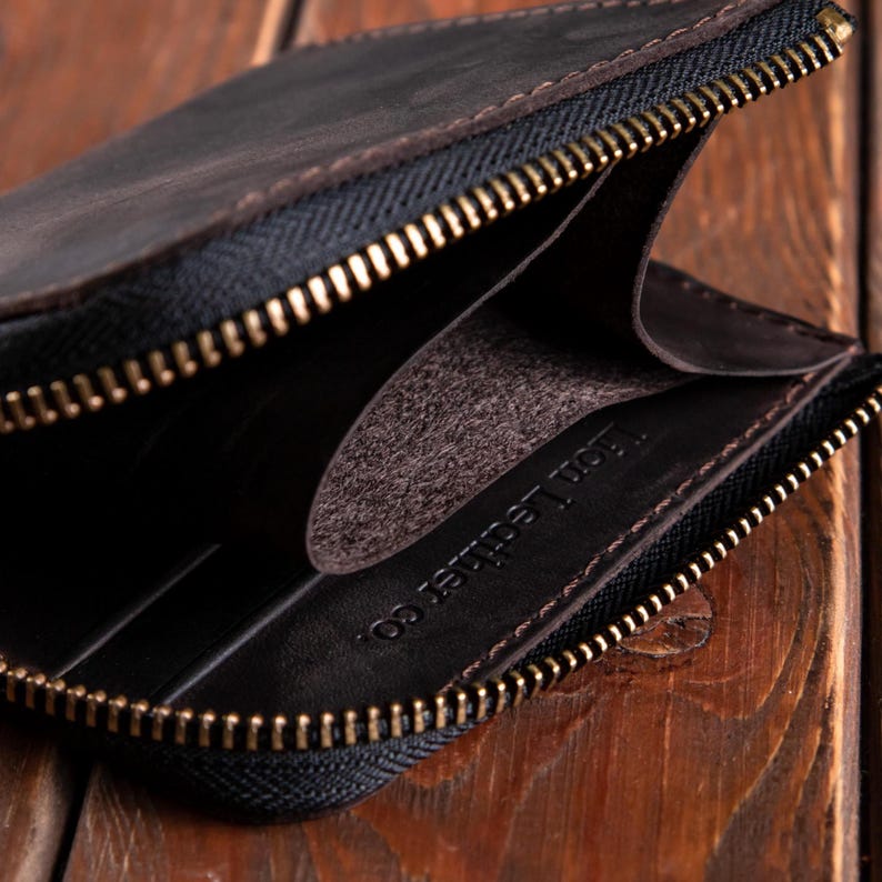 May include: A dark brown leather wallet featuring a brass-coloured metal zip closure. The interior is lined with dark brown leather and displays the embossed text Lion Leather co. on the inner side panel, showing a single open compartment.