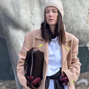 May include: A beige wool coat with neon yellow stitching details, worn over a white hooded sweatshirt. A dark brown rectangular handbag with a silver zip and a matching shoulder strap is held, alongside dark red leather gloves.