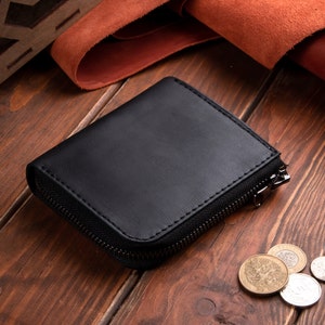 May include: A black leather wallet with a smooth matte finish and a dark metal zip closure. The wallet is rectangular with rounded corners and visible stitching along the edges. Several metallic coins are scattered near the wallet.
