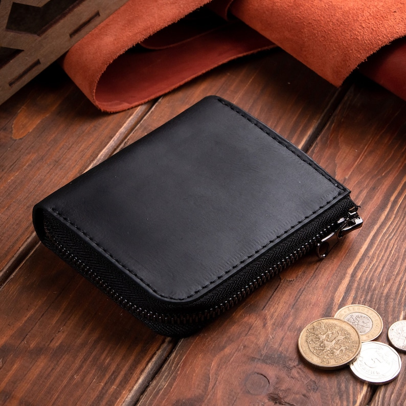 May include: A black leather wallet with a smooth matte finish and a dark metal zip closure. The wallet is rectangular with rounded corners and visible stitching along the edges. Several metallic coins are scattered near the wallet.