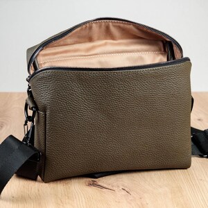 May include: An olive green crossbody bag with a textured pebble grain finish, featuring a black zipper closure along the top, a beige fabric interior lining, and a black adjustable nylon strap attached with black metal hardware clips on the sides.