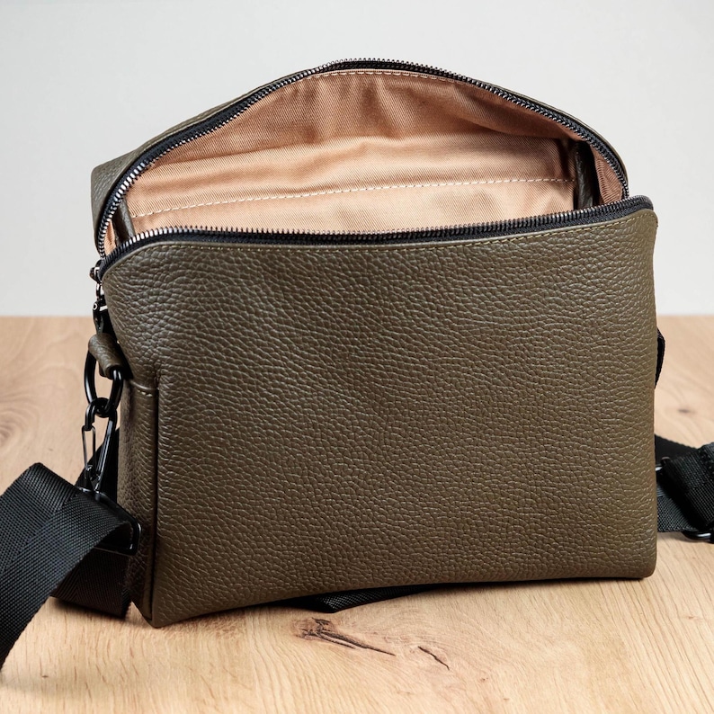 May include: An olive green crossbody bag with a textured pebble grain finish, featuring a black zipper closure along the top, a beige fabric interior lining, and a black adjustable nylon strap attached with black metal hardware clips on the sides.