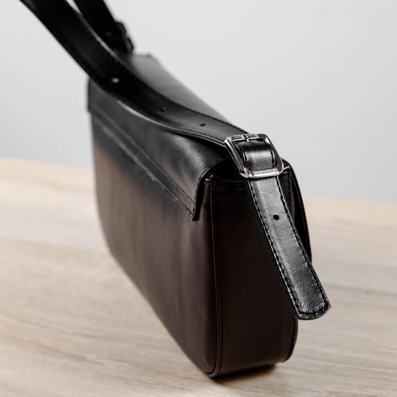 Op de afbeelding: Een zwarte schoudertas met een gladde afwerking, voorzien van een lange verstelbare riem die vastzit met een zilverkleurige metalen gesp. De tas heeft een gestructureerde rechthoekige vorm met afgeronde hoeken aan de onderkant.