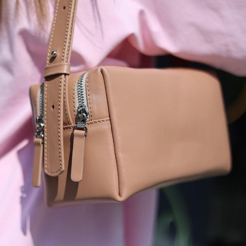 Pu&ograve; includere: Una borsa a tracolla rettangolare in pelle beige con due cerniere in metallo argentato e tiralampo in pelle abbinati. La borsa include un'ampia tracolla in pelle regolabile fissata con un rivetto in metallo argentato.