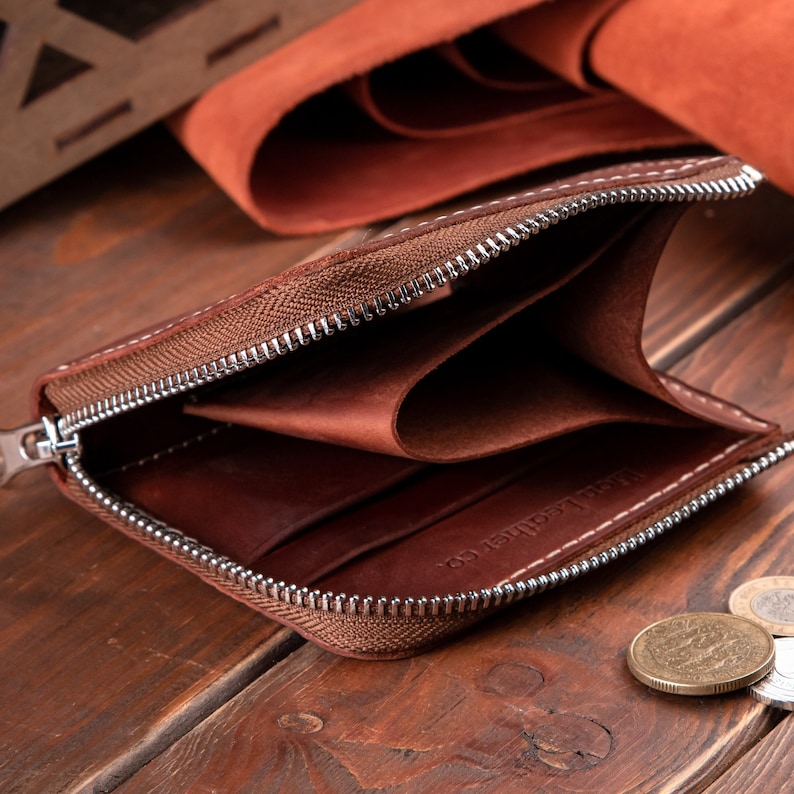 May include: A brown leather wallet with a silver metal zip closure. The interior features multiple card slots and a main compartment. The text "Men Leather Co." is embossed on the inner surface. The wallet is open, revealing its smooth texture.