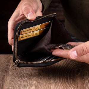 May include: A black zipped wallet with a dark brown interior lining. The wallet features multiple internal slots containing gold-coloured credit cards and a central compartment holding several silver-toned metal coins. The zip is metallic.