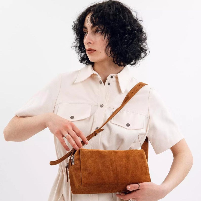 Pu&ograve; includere: Una borsa a tracolla rettangolare in pelle scamosciata color marrone tabacco con chiusura a zip superiore e una sottile tracolla regolabile. La borsa presenta una silhouette strutturata e squadrata con cuciture visibili e una zip argentata.