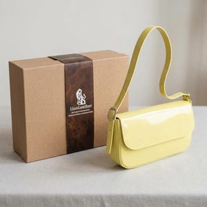 Può includere: Una borsa a tracolla giallo pallido lucida con un unico cinturino regolabile e una fibbia color argento, posizionata accanto a una scatola regalo rettangolare in cartone con un'etichetta marrone scuro con il logo e il testo LionLeather.
