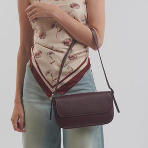 Può includere: Una borsa a tracolla rettangolare color bordeaux scuro con finitura liscia, patta ripiegabile e un cinturino sottile regolabile. La borsa è indossata con un top a foulard color crema con motivo floreale rosso e jeans azzurri.
