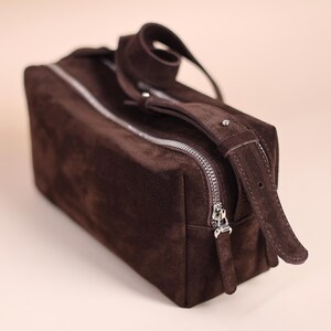 May include: A rectangular dark brown suede wash bag featuring a silver-toned metal zip closure that runs along the top. The bag includes a matching suede handle attached to the side with a small silver metal stud detail for secure carrying.
