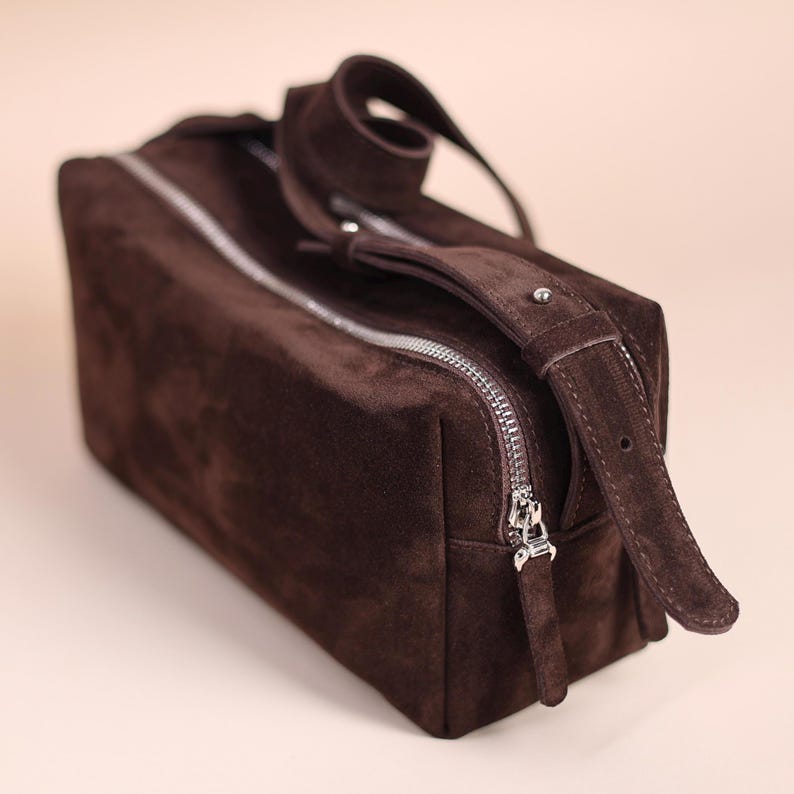 May include: A rectangular dark brown suede wash bag featuring a silver-toned metal zip closure that runs along the top. The bag includes a matching suede handle attached to the side with a small silver metal stud detail for secure carrying.