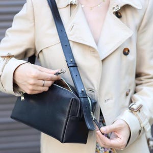 Pu&ograve; includere: Una borsa a tracolla in pelle nera di forma rettangolare, con cerniere in metallo argentato e una tracolla regolabile. La borsa &egrave; indossata con un trench beige con bottoni scuri e colletto.