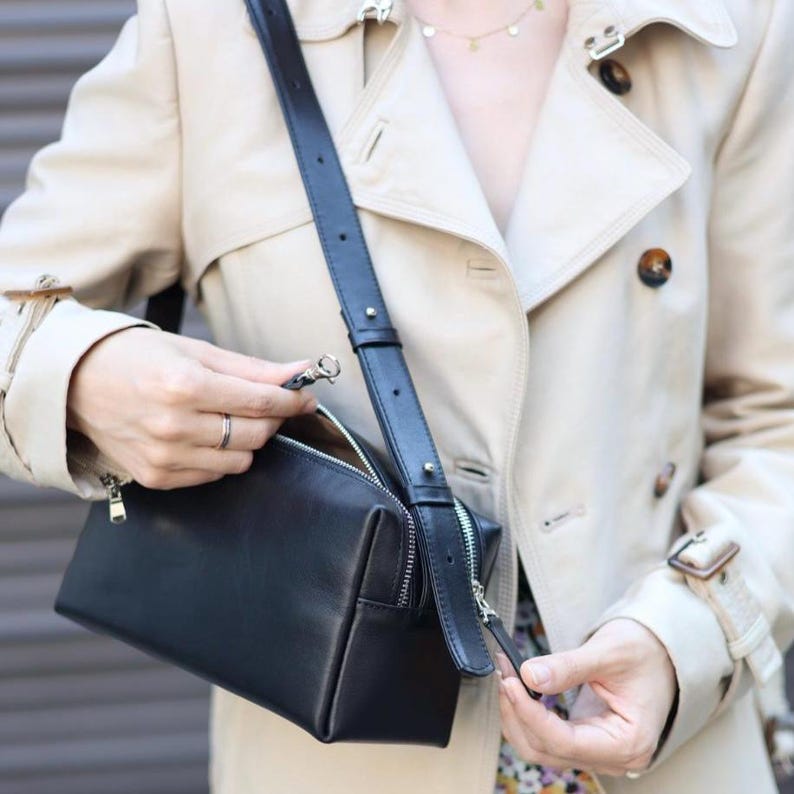 Pu&ograve; includere: Una borsa a tracolla in pelle nera di forma rettangolare, con cerniere in metallo argentato e una tracolla regolabile. La borsa &egrave; indossata con un trench beige con bottoni scuri e colletto.
