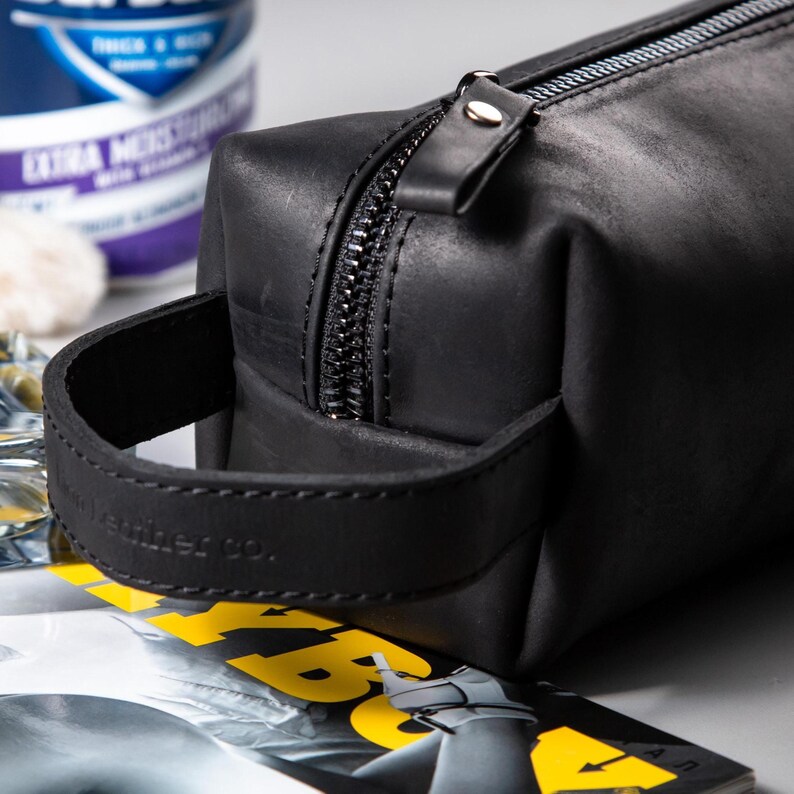 May include: A matte black leather wash bag featuring a rectangular shape, a sturdy side handle with embossed text, and a black metal zip closure. The bag has a smooth, dark finish and a structured design with a pull tab on the zip.