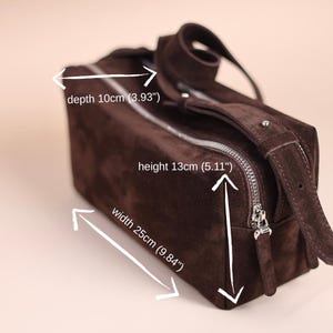 Pu&ograve; includere: Una borsa rettangolare in camoscio marrone scuro con chiusura a zip argentata e tracolla regolabile. La borsa misura 25 cm di larghezza, 13 cm di altezza e 10 cm di profondit&agrave;, come indicato dal testo e dalle frecce bianche nell'immagine.
