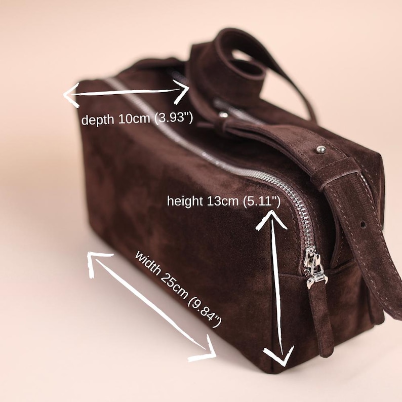 Pu&ograve; includere: Una borsa rettangolare in camoscio marrone scuro con chiusura a zip argentata e tracolla regolabile. La borsa misura 25 cm di larghezza, 13 cm di altezza e 10 cm di profondit&agrave;, come indicato dal testo e dalle frecce bianche nell'immagine.