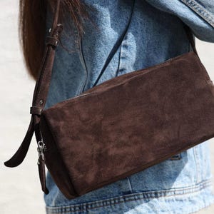 Pu&ograve; includere: Una borsa a tracolla in camoscio marrone scuro dalla silhouette rettangolare, dotata di una tracolla regolabile abbinata con fibbia in metallo e una chiusura a zip color argento con una lunga linguetta in camoscio, indossata sopra una giacca di jeans blu.