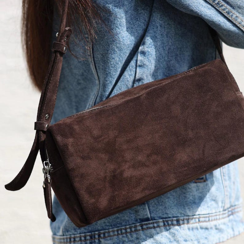 Pu&ograve; includere: Una borsa a tracolla in camoscio marrone scuro dalla silhouette rettangolare, dotata di una tracolla regolabile abbinata con fibbia in metallo e una chiusura a zip color argento con una lunga linguetta in camoscio, indossata sopra una giacca di jeans blu.