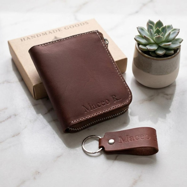 May include: A brown leather bi-fold wallet with a zip closure and the name Maceo R. embossed on the front, accompanied by a matching brown leather keyring with the name Maceo embossed, both resting near a cardboard box and a small succulent.