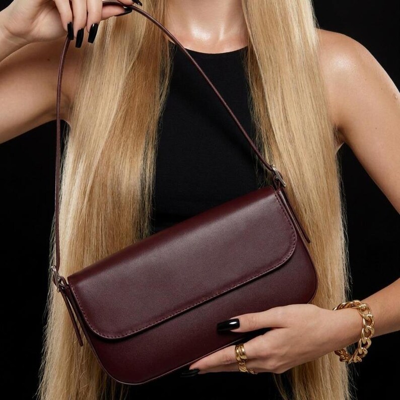 May include: A deep burgundy rectangular shoulder bag with a smooth finish, a fold-over flap closure, and a thin matching shoulder strap. The bag features clean lines and a minimalist structure, providing a sleek and functional accessory for daily use.