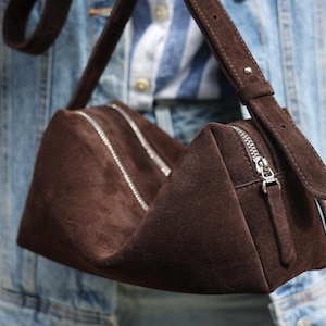 May include: A dark brown suede handbag with a structured, triangular silhouette. The bag features two silver-toned metal zips running along the top, a matching suede shoulder strap with a metal buckle, and a silver zip pull on the side.