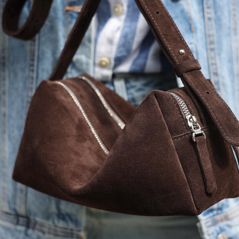 May include: A dark brown suede handbag with a structured, triangular silhouette. The bag features two silver-toned metal zips running along the top, a matching suede shoulder strap with a metal buckle, and a silver zip pull on the side.