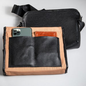 May include: A black pebbled leather crossbody bag with a black nylon strap and a separate tan fabric insert featuring two black leather exterior pockets. A smartphone and a small brown leather card holder are tucked into the front pockets.