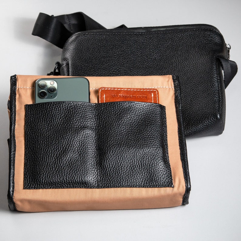 May include: A black pebbled leather crossbody bag with a black nylon strap and a separate tan fabric insert featuring two black leather exterior pockets. A smartphone and a small brown leather card holder are tucked into the front pockets.