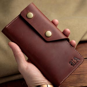 Może przedstawiać: Brązowy skórzany portfel z klapką zabezpieczoną dwoma zatrzaskami w kolorze mosiądzu. Portfel ma białe przeszycia wzdłuż krawędzi, a inicjały "LA" są widoczne.