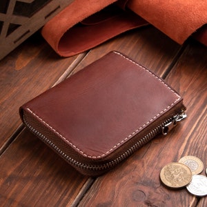 May include: A dark brown leather wallet with a rectangular shape and rounded corners. The wallet features visible white stitching along the edges and a silver-toned metal zip that runs along three sides of the exterior, securing the contents inside.