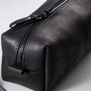 May include: A rectangular black leather pouch featuring a matte finish and a black zip closure. The top of the zip includes a matching black leather pull tab secured with a small silver-toned metal rivet, providing a functional opening mechanism.