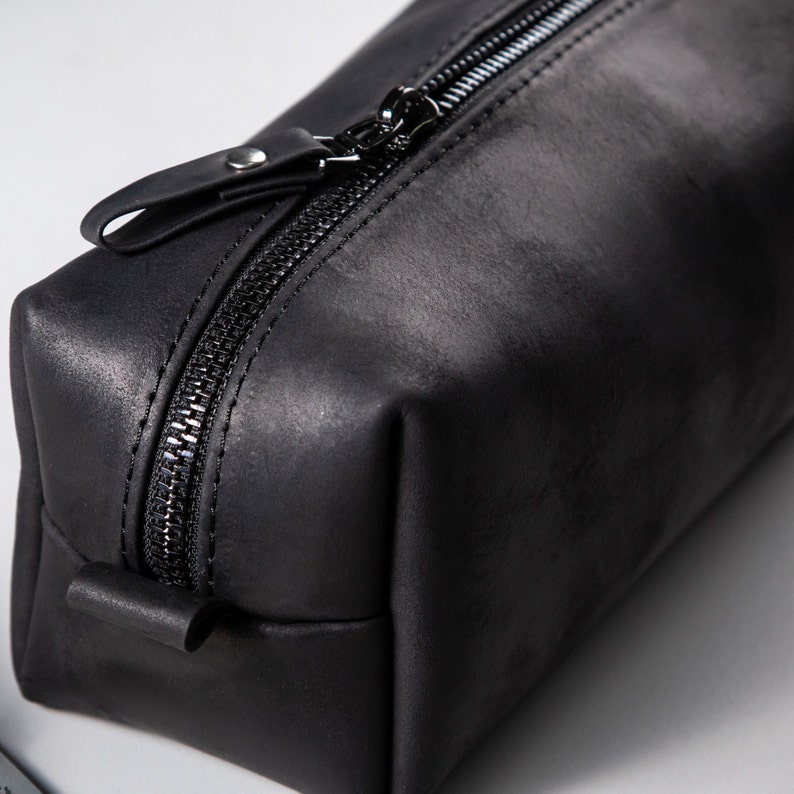 May include: A rectangular black leather pouch featuring a matte finish and a black zip closure. The top of the zip includes a matching black leather pull tab secured with a small silver-toned metal rivet, providing a functional opening mechanism.