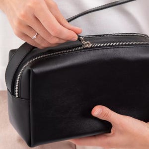 Pu&ograve; includere: Una trousse rettangolare nera con finitura liscia e opaca e chiusura a zip in metallo color argento. La borsa presenta un design strutturato con linee pulite e un unico scomparto principale, offrendo una soluzione di archiviazione semplice e funzionale.