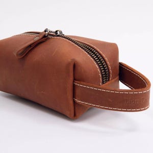 May include: A brown leather toiletry bag featuring a top zip closure with a metal pull tab and a side handle strap. The bag is constructed with visible white stitching along the edges and includes the embossed text Lion Leather on the handle.