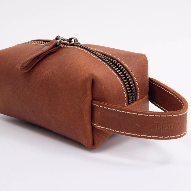 May include: A brown leather toiletry bag featuring a top zip closure with a metal pull tab and a side handle strap. The bag is constructed with visible white stitching along the edges and includes the embossed text Lion Leather on the handle.