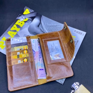 Puede incluir: Una cartera larga de cuero marr&oacute;n con m&uacute;ltiples ranuras para tarjetas, una ventana transparente para identificaci&oacute;n y un compartimento con cremallera. La cartera est&aacute; abierta, mostrando varias tarjetas de cr&eacute;dito doradas y billetes en su interior.