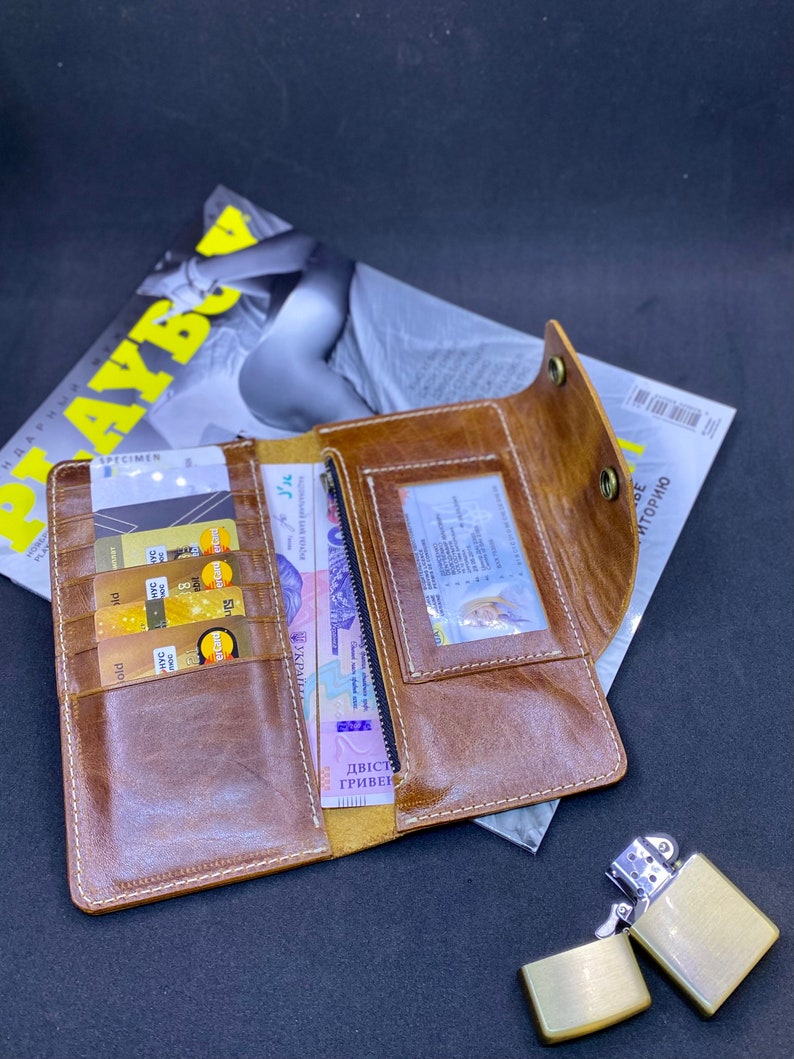 Puede incluir: Una cartera larga de cuero marr&oacute;n con m&uacute;ltiples ranuras para tarjetas, una ventana transparente para identificaci&oacute;n y un compartimento con cremallera. La cartera est&aacute; abierta, mostrando varias tarjetas de cr&eacute;dito doradas y billetes en su interior.