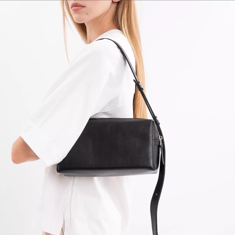 Pu&ograve; includere: Una borsa a tracolla rettangolare nera con finitura liscia, dotata di un cinturino sottile regolabile e una chiusura a zip in metallo color argento sul bordo superiore, indossata sopra una maglietta bianca.