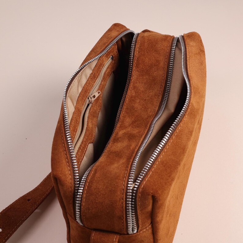 Pu&ograve; includere: Una borsa a tracolla in camoscio marrone con due scomparti principali con cerniera e una tasca interna pi&ugrave; piccola con cerniera. La borsa presenta cerniere in metallo color argento e una fodera in tessuto beige chiaro visibile all'interno.